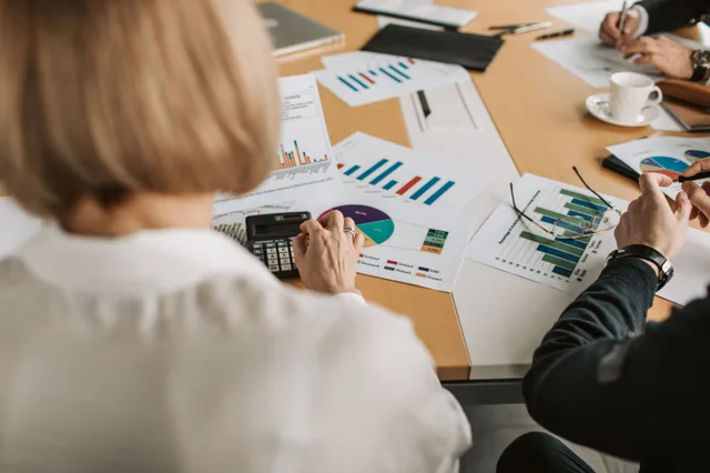 Overhead view of professionals discussing financial graphs and using calculators in an office setting. Omnichannel Digital Planning Agency Fee Models